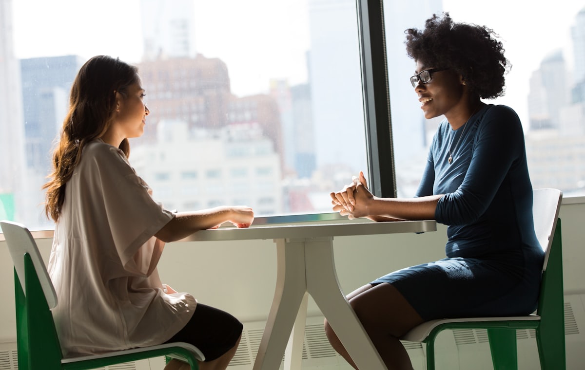 Two people sitting across a table from each other, mid-conversation,
            in a room with a large window.